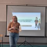 Kobieta o jasnych włosach, ubrana w brązowy sweter i jeansy. Stoi na tle ekranu projekcyjnego, na którym widnieje napis "Nie muszę atakować, żeby się bronić".