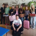 Grupa uśmiechniętej młodzieży w sali lekcyjnej podczas szkolnego wydarzenia "Dzień bez plecaka". Zamiast tradycyjnych plecaków, uczniowie trzymają nietypowe przedmioty na książki: walizka, chlebak, koszyk, itp.