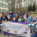 Grupa dzieci i dorosłych zgromadzonych na zewnątrz podczas marszu. Dwie kobiety trzymają baner z napisem " Publiczna Szkoła Podstawowa z Oddziałami Integracyjnymi i Sportowymi Nr 5 im. Jana Pawła II w Pionkach".