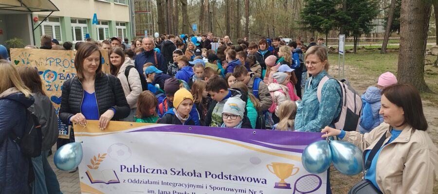 Grupa dzieci i dorosłych zgromadzonych na zewnątrz podczas marszu. Dwie kobiety trzymają baner z napisem " Publiczna Szkoła Podstawowa z Oddziałami Integracyjnymi i Sportowymi Nr 5 im. Jana Pawła II w Pionkach".