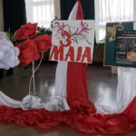 Patriotyczna dekoracja z okazji Święta 3 Maja. W środku znajduje się biała plansza z wyraźnym napisem "S MAJA". w kolorze czerwonym, ozdobiona subtelnym motywem kwiatowym. Cała kompozycja bazuje na bieli i czerwieni. Po lewej duże dekoracyjne białe kwiaty, po prawej analogiczne kwiaty czerwone.