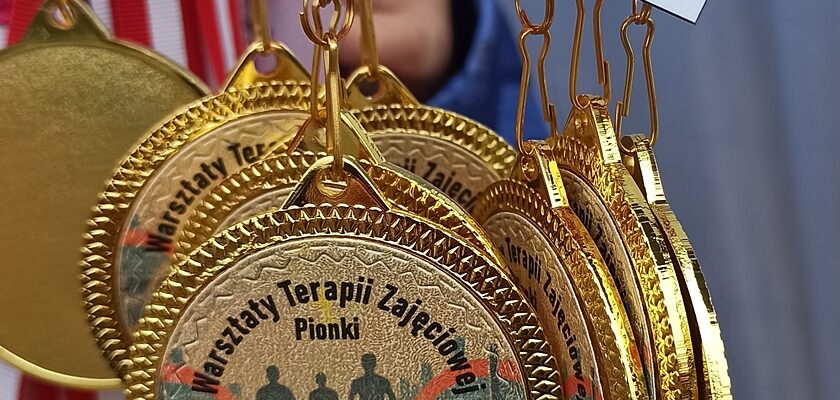 Pamiątkowe medale sportowe wiszące na biało-czerwonych wstążkach. W centrum medalu znajduje się naklejka z napisem "Warsztaty Terapii Zajęciowej Pionki XIX impreza sportowa".