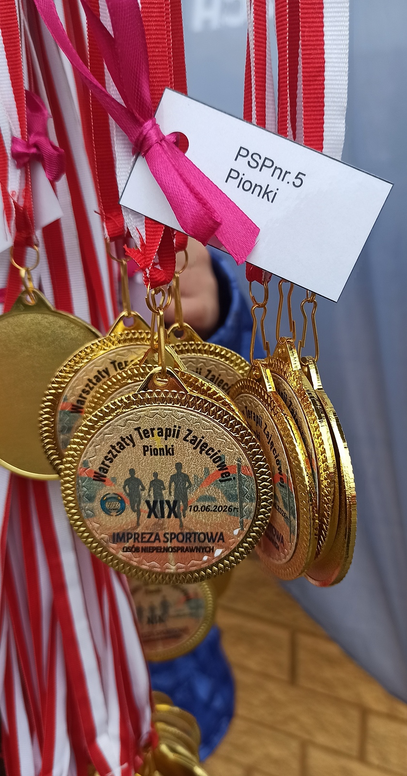 Pamiątkowe medale sportowe wiszące na biało-czerwonych wstążkach. W centrum medalu znajduje się naklejka z napisem "Warsztaty Terapii Zajęciowej Pionki XIX impreza sportowa".