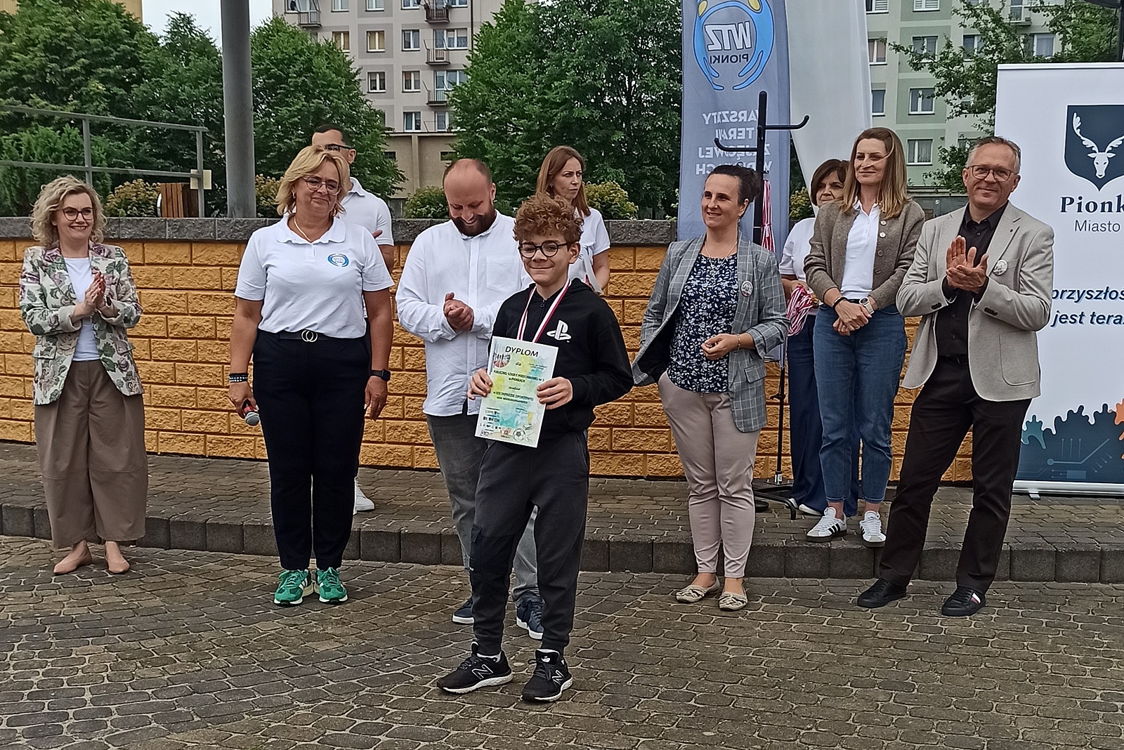 Moment wręczania nagrody podczas wydarzenia plenerowego. Na środku stoi uśmiechnięty chłopiec. W rękach trzyma pamiątkowy dyplom, a na szyi ma zawieszony medal.