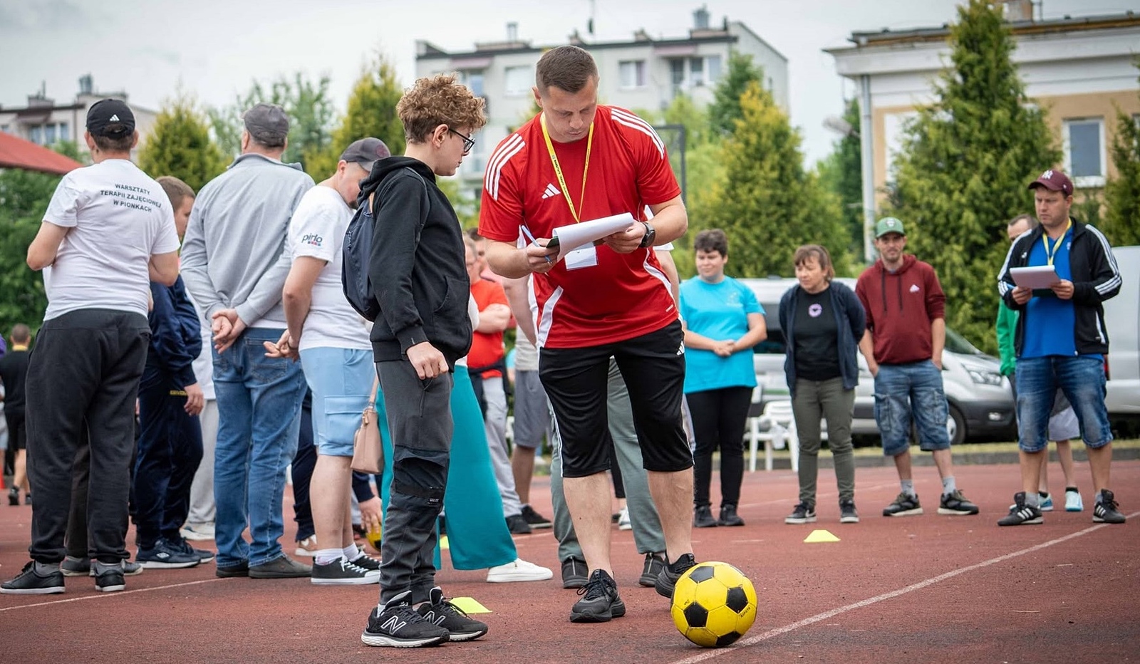 Mężczyzna w czerwonej koszulce sportowej pochyla się, pisząc coś na trzymanej w rękach kartce papieru. Obok stoi nastoletni chłopiec. W tle grupa dorosłych.