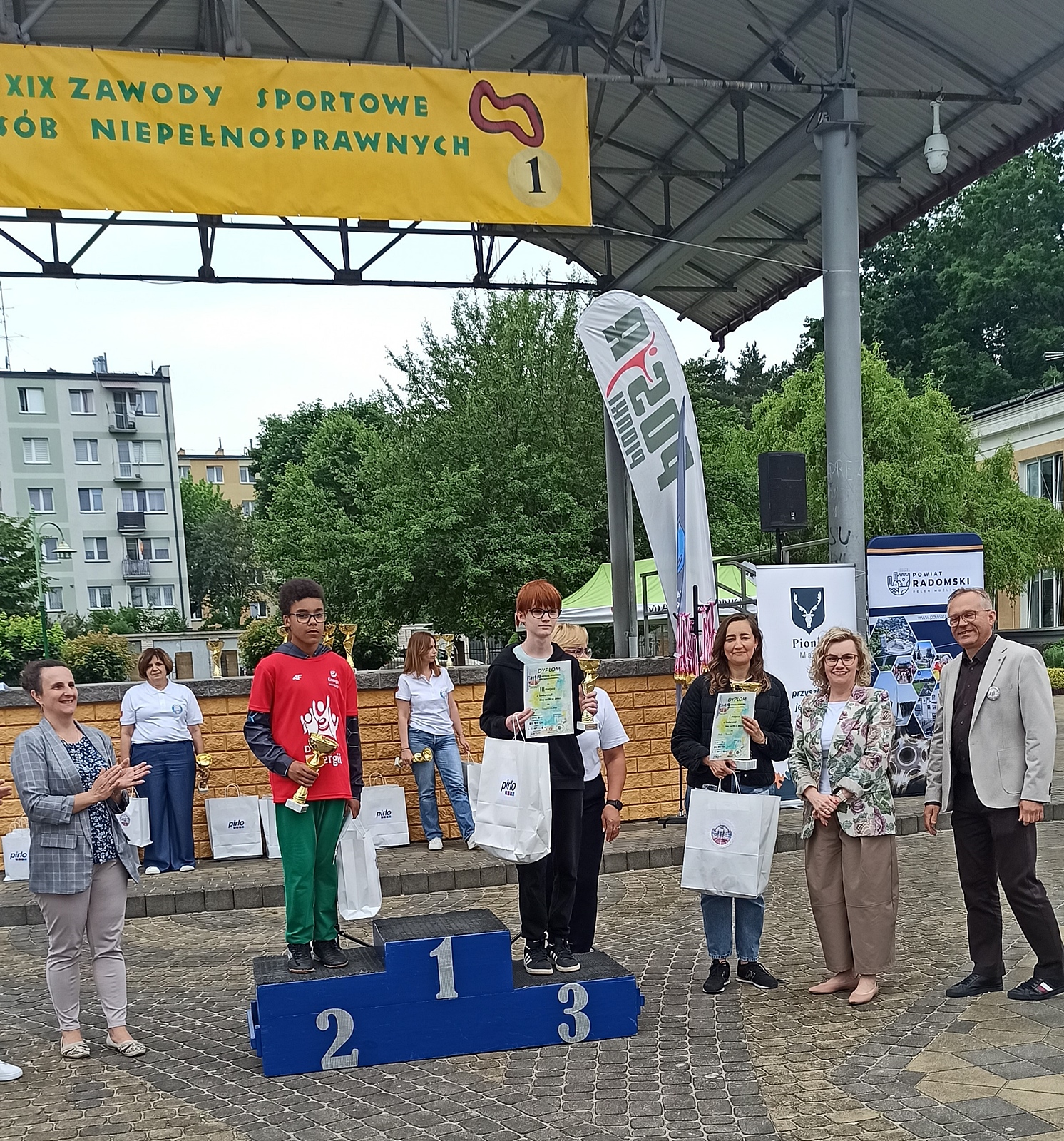 Ceremonia wręczania nagród na scenie plenerowej. Nad sceną wisi żółty baner z czarnym napisem" XIX zawody sportowe osób niepełnosprawnych".