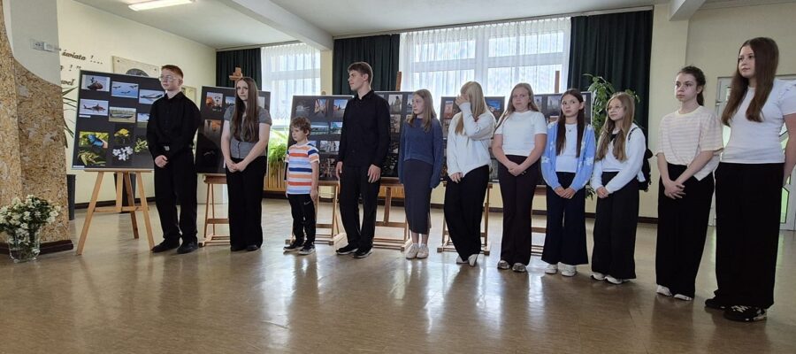 Grupa dzieci i młodzieży stojąca w rzędzie na tle wystawy prac fotograficznych.