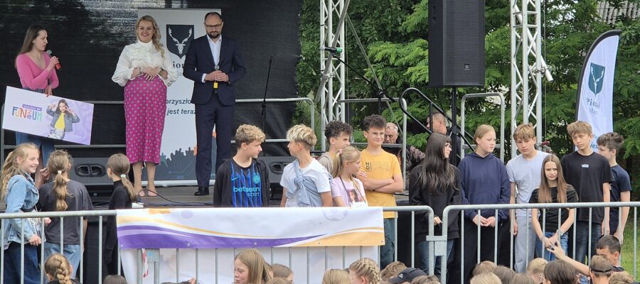 Na plenerowej scenie stoją trzy dorosłe osoby - dwie kobiety i jeden mężczyzna trzymający mikrofon. Przed sceną stoi grupa nastolatków.