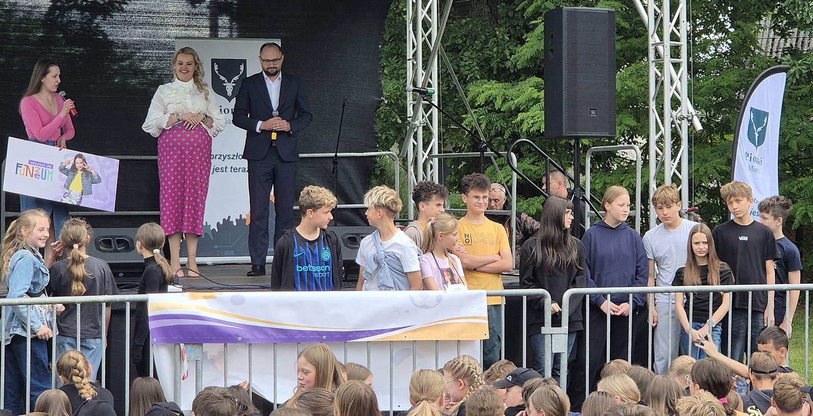 Na plenerowej scenie stoją trzy dorosłe osoby - dwie kobiety i jeden mężczyzna trzymający mikrofon. Przed sceną stoi grupa nastolatków.