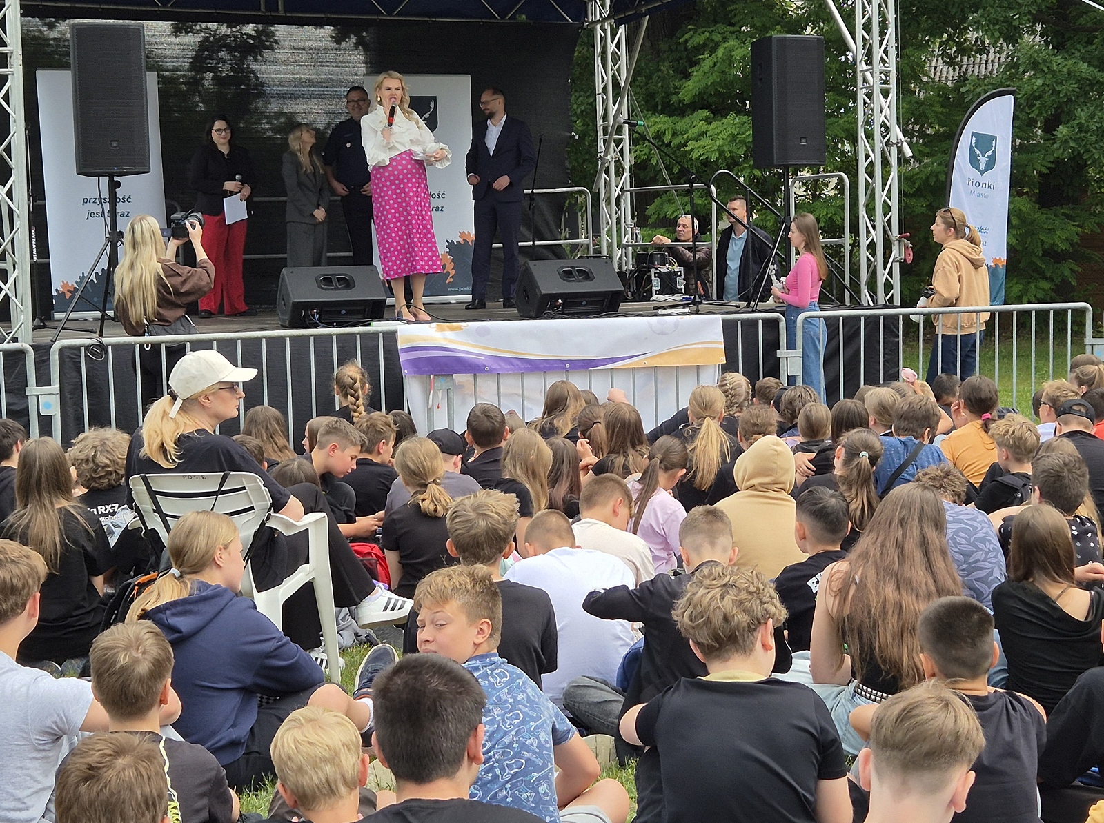 Na plenerowej scenie stoi centralnie kobieta w białej bluzce i różowej spódnicy w groszki, która mówi do mikrofonu. Przed sceną siedzi na ziemi oraz na krzesełkach liczna grupa dzieci i młodzieży sfotografowana od tyłu.