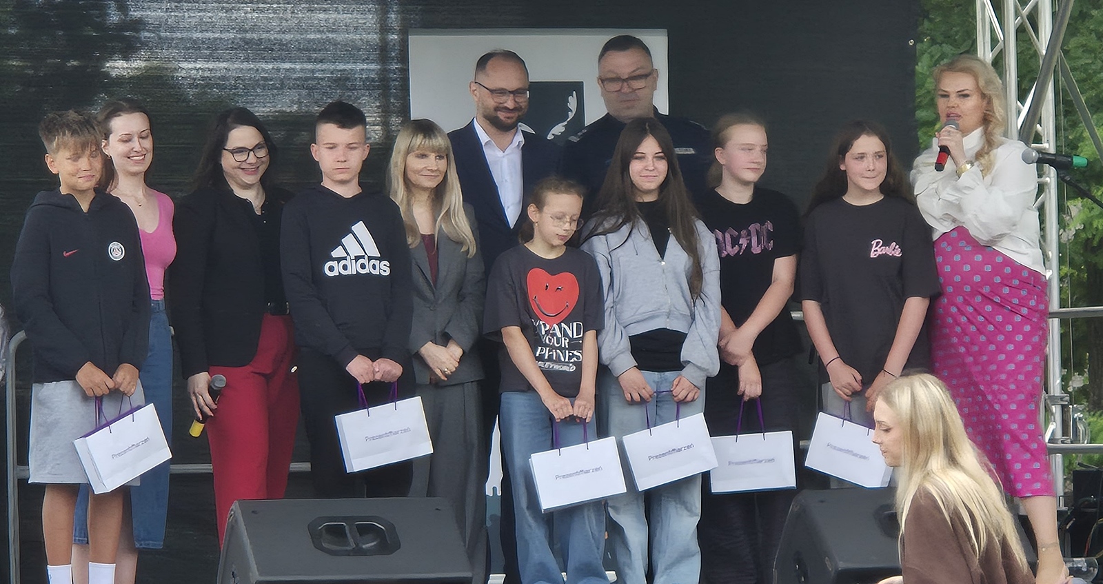 Grupa dzieci, młodzieży i dorosłych stojąca na plenerowej scenie. Młodzi ludzie trzymają w rękach białe, upominkowe torebki.