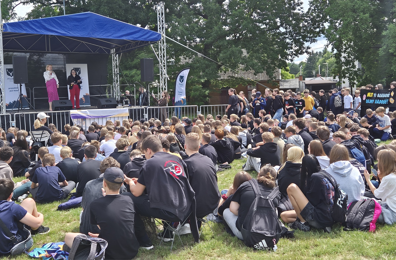 Kadr z dalszej perspektywy pokazuje liczną grupę dzieci i młodzieży. Siedzą tyłem do obiektywu, bezpośrednio na trawie przed sceną. W głębi znajduje się plenerowa scena zna której znajdują się dwie dorosłe kobiety.