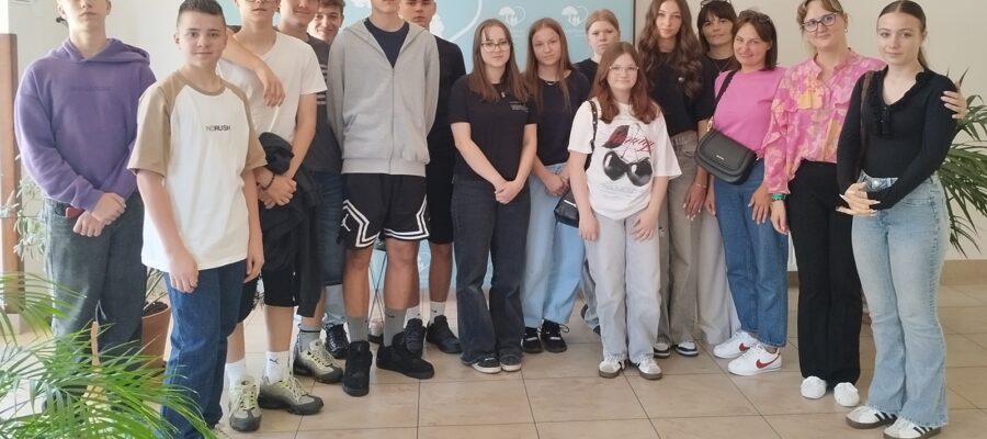 Grupa młodzieży i dorosłych stojących obok siebie. Grupa stoi w jasnym pomieszczeniu na tle ściany, na której widać fragment zielonej grafiki.