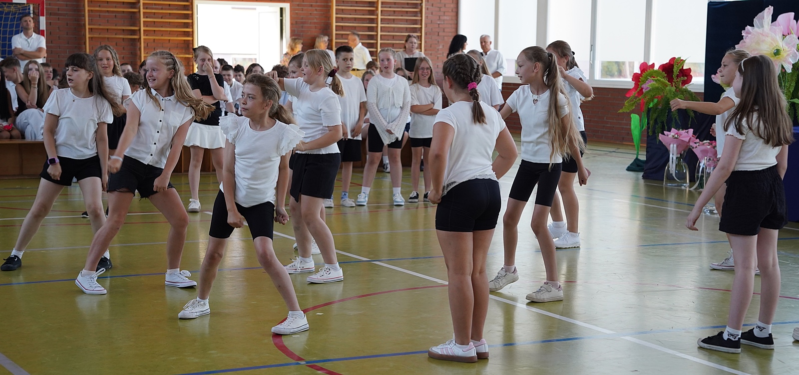 Grupa dzieci podczas występu taneczno-sportowego na uroczystości szkolnej. Dziewczynki w wieku wczesnoszkolnym, uchwycone w dynamicznych, tanecznych pozach.