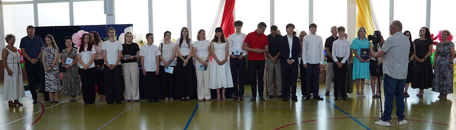 Grupa nastolatków wraz z rodzicami stojących w rzędzie podczas uroczystości szkolnej.