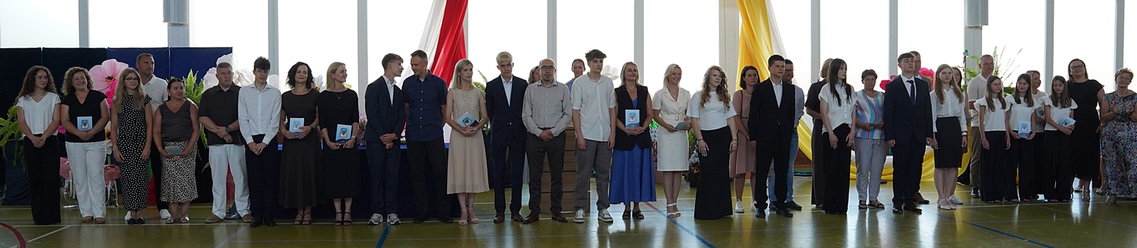 Grupa nastolatków wraz z rodzicami stojących w rzędzie podczas uroczystości szkolnej.